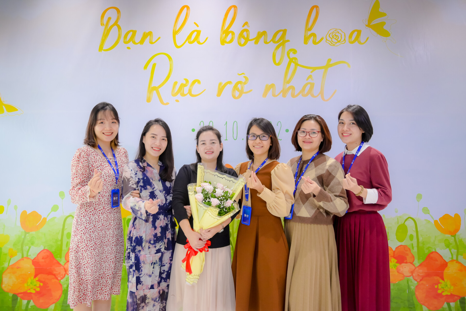 chị em các phòng ban tại VPHN xúng xính váy áo dưới backdrop "Bạn là bông hoa rực rỡ nhất"